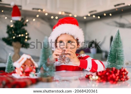 Cute boy kid in a red Santa hat drinking filtered water from a glass in the kitchen. Holidays, health concept.