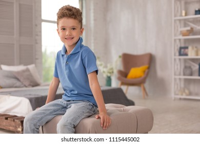 Cute Boy. Little Pleasant Looking Cute Boy Wearing Dark Blue Shirt And Denim Jeans Sitting On Hassock In The Living Room