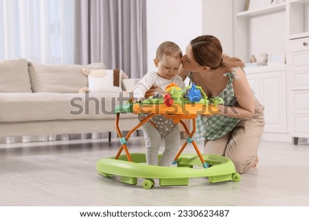 Cute boy making first steps with baby walker. Mother kissing her little son at home, space for text