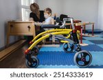 Cute boy with neurodevelopmental disorders is participating in a rehabilitation session under guidance of therapist in therapy space with rehab walker