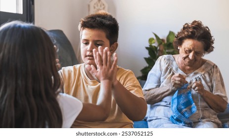 Cute boy playing clapping game with sister in living room while grandmother knitting sweater in background at home - Powered by Shutterstock