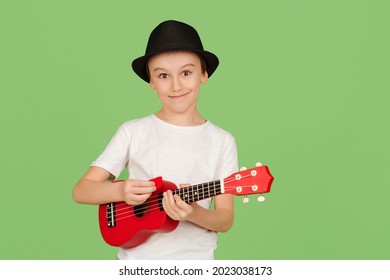 Cute Boy Plays The Ukulele. Happy Kid Enjoying The Music. Student Learning To Play Ukuleles. Fashionable Boy In Summer Hat Isolated Over Green Background.