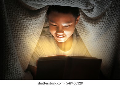 Cute boy reading book under blanket at night. Magic light coming out of book.