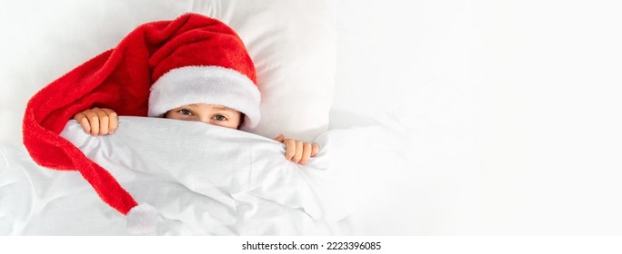 A Cute Boy In A Red Santa Hat Lies In A Bed In The Bedroom And Looks Out From Under The Covers. Joyful Child Hides In A Blanket. Christmas Morning Background With Copy Space For Text.