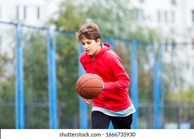 Cute Boy In Red T Shirt Plays Basketball On City Playground. Active Teen Enjoying Outdoor Game With Orange Ball. Hobby, Active Lifestyle, Sport For Kids.