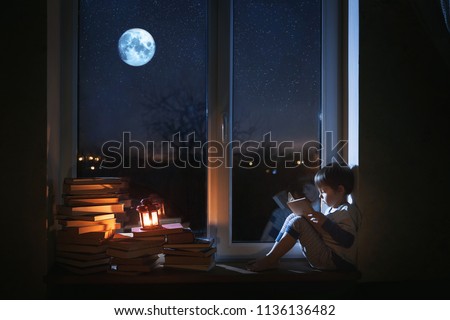 A cute boy sits on the windowsill at night. The child reads books under the moonlight. The window shows the moon and stars.