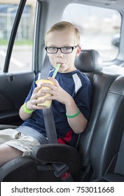 Cute Boy Sitting In A Booster Seat On A Long Car Ride Drinking A Smoothie While Safely Strapped In A Car Seat. He Is Wearing His Seatbelt And Enjoying A Drink.