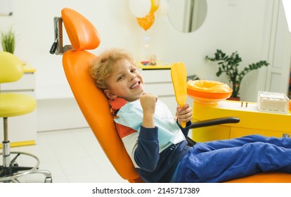 A Cute Boy Is Sitting In A Dental Chair In A Modern Clinic. A Charming Baby Looks Into A Stamological Mirror. The Concept Of Pediatric Dentistry