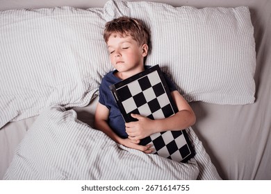Cute boy sleeping in bed while hugging a chessboard, dreaming about strategy, intelligence and childhood imagination. Concept of passion for chess, learning,peaceful rest. Future chess grandmaster.