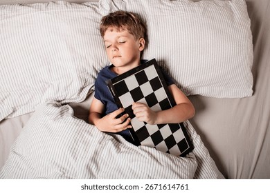 Cute boy sleeping in bed while hugging a chessboard, dreaming about strategy, intelligence and childhood imagination. Concept of passion for chess, learning,peaceful rest. Future chess grandmaster.