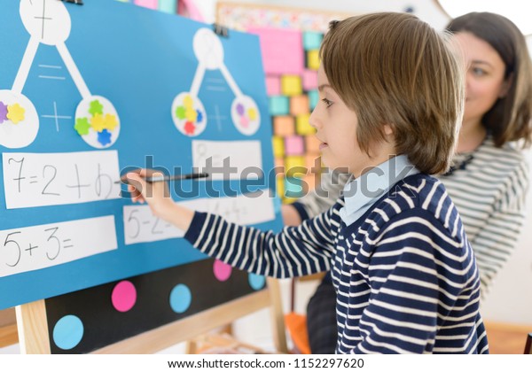 Cute boy solving graphic math exercises at\
kindergarten, under educator\
observation