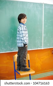 Cute Boy Standing On A Chair In Front Of Blackboard