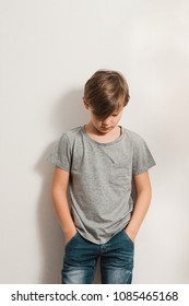a cute boy stands next to white wall, grey t-shirt, blue jeans, hands in pockets, bowing his head down