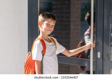 Cute Boy Student With A School Backpack Opens The Door Of The House. A Child Returning Home After School Next To The Door Of The House. Back To School Concept.