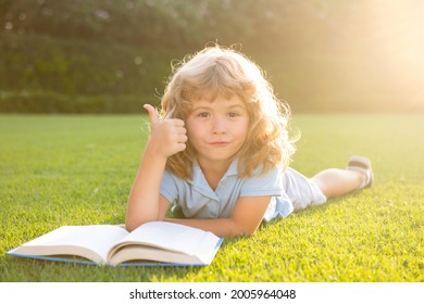 Cute Boy With Thumbs Up Reading Book On Green Grass. Summer Outdoor Kids Education.