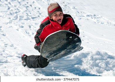 Cute Boy In Tobaggan Riding Down A Sled On A Snow Hill With Tongue Sticking Out.