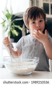 A Cute Boy, A Toddler Of Caucasian Appearance, Cooks In The Kitchen And Tastes The Dough. Vertical Photo. Ideal For Illustrating Real Life Moments