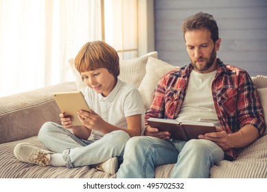 Cute boy is using a digital tablet and smiling while his handsome father is reading a book, both are sitting on couch at home