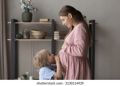 Cute boy using medical stethoscope, listening to pregnant mom big tummy, asking about pregnancy, childbirth, expected younger sibling. Expecting mother having fun with kid