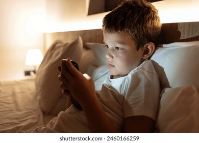 Cute boy using a smartphone. Child playing with mobile phone, lying on a bed. Technology and internet concept. Boy laying on bed and looking at smartphone. Light reflection on kid face from phone - Powered by Shutterstock