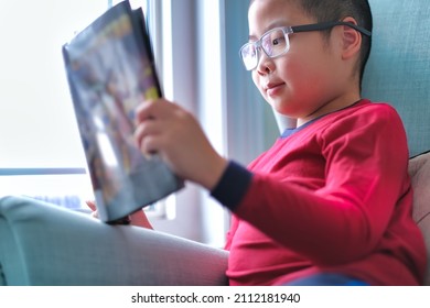 Cute Boy Wearing Glasses, Smiling And Reading A Fun Graphic Comic Novel Book Near Window At Home. Child Learning Activity Background.