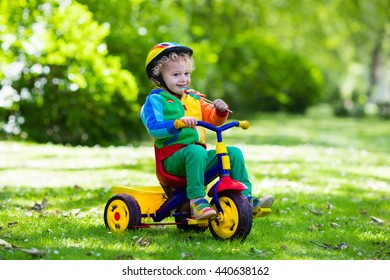 Cute Boy Wearing Safety Helmet Riding His Tricycle In Sunny Summer Park. Kids Ride Bicycle. First Bike For Little Child. Active Toddler Kid Playing And Cycling Outdoors. Kids Play In The Garden.