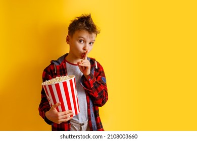 Cute Boy Who Is Asking To Be Quiet With Finger On Lips With Huge Popcorn Bucket On Yellow Background. Kid Is Wearing Casual Plaid Shirts Outfit Isolated. Movie Concept. Teenager Showing Quiet Gesture