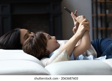 Cute Boy And Young Mom Using Smartphone, Resting On Couch Together, Taking Home Selfie, Texting, Chatting Online, Looking At Screen. Happy Mother And Kid Holding Mobile Phone, Relaxing On Sofa