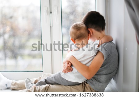 cute boys on the windowsill by window, older brother hugs younger, taking care of sibling. concept siblings and candid lifestyle