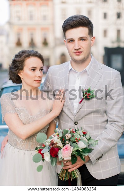 Cute bride in long\
wedding dress and groom, young newlyweds, just married couple,\
standing on streets of old city center on wedding day near black\
retro car outdoors.