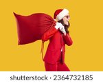 Cute brunette with beautiful hair in sunglasses, dressed in a business red Santa Claus suit carries a bag of gifts. Beautiful girl carries a large red bag with gifts on an isolated yellow background.
