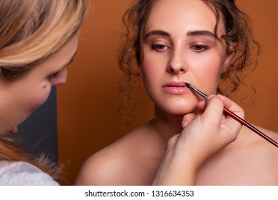 Cute Brunette Caucasian Woman With Fresh, Clean, Healthy, Flawless Skin. Nude, Day Make Up With Casual Hairstyle. Portrait On A Brown Background. Backstage Of Make Up Artist Applying Lipstick On Model