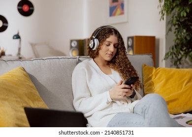 Cute Brunette Dressed In A White Sweater And Blue Jeans Listens To Her Favorite Music Through Wireless Headphones, Holding A Phone.