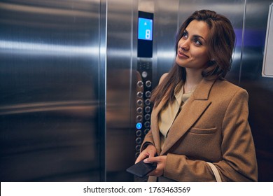Cute brunette female in trendy coat staying in lift alone with mobile in hands and smiling