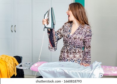 Cute, Brunette Girl Ironing Shirt On Ironing Board In Her Bedroom. Funny Expression, Iron Is Too Hot. Housekeeping Concept