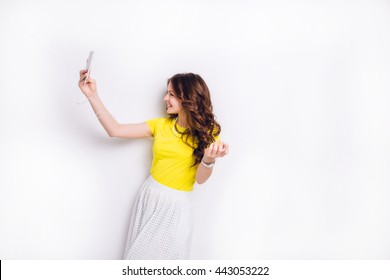 Cute brunette girl listening to music on earphones on smartphone is having fun and takes a selfie. She smiles widely. She has long wavy brunette hair. She wears yellow t-shirt and white skirt.