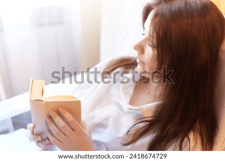 Cute brunette girl is reading small book of poetry in bedroom in morning. Young pregnant woman in the maternity hospital with a book of tips on parenting. A sick and recovering lady