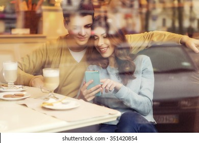 Cute Brunette Loving Couple In Cozy Warm Sweaters On A Date At The Cafe Looking At The Phone, Using It. Cold Autumn, Winter Or Spring Day, European City. Indoors