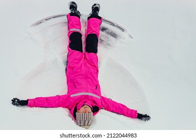 Cute Brunette Teen Girl In Bright Clothing Lying On Snow And Making Snow Angel Figure With Hands And Legs. Winter Outdoor Fun