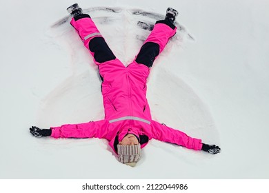 Cute Brunette Teen Girl In Bright Clothing Lying On Snow And Making Snow Angel Figure With Hands And Legs. Winter Outdoor Fun