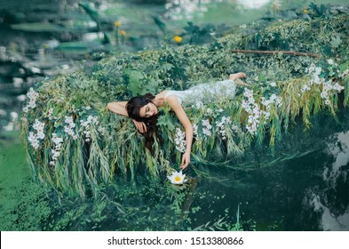 Cute Brunette Woman Long Hair Lies In Boat  Gently Touches Water Lily Her Hand. Princess Bride Dress Floats On Lake. Friday, Sleeping Relaxation Enjoyment. Creative Wedding Decor Natural Cosmetics 