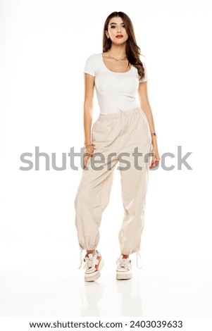 Cute brunette woman in white shirt and loose white trousers posing on a white studio background. Full length, front view.