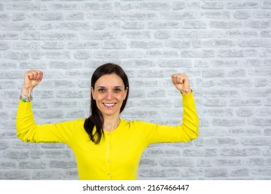 Cute Brunette Woman In Yellow Sweater, With On Gray Background. Strong Girl Shows Muscles, Bent Arm At The Elbow. Happy Strong  Woman Show Biceps Hands Winner Isolated On Grey Brick Wall Background