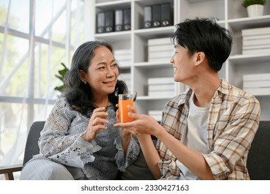 A cute and caring Asian grandson is giving a glass of orange juice to his grandmother, spending quality time at home together. family bonding, happy family, love and care - Powered by Shutterstock