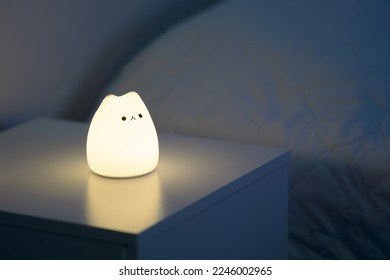 Cute cat shaped night lamp standing on a bedside table next to bed. Bedside lamp. Night lamp standing next to bed. Bedroom lamp on a night table next to a sleeping bed in a dark room. Close up. 