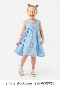 A cute Caucasian 5-year-old girl with a beautiful smile with a fashionable hairstyle stands on a white background in full height in a blue summer sundress and laughs with narrowed eyes.