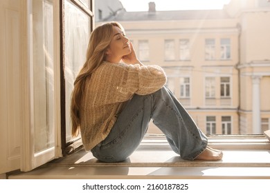 Cute caucasian adult lady with her eyes closed is basking in sun sitting on windowsill on warm day. Long-haired blonde wears casual clothes. Concept of enjoying moment - Shutterstock ID 2160187825