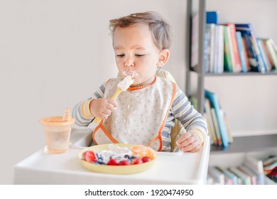 Cute Caucasian Baby Boy Eating Ripe Berries And Fruits With Yogurt. Funny Smiling Child Kid Sitting In Chair Eating Fresh Berries And Drinking Water. Supplementary Healthy Food For Toddlers Kids. 