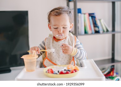 Cute Caucasian Baby Boy Eating Ripe Berries And Fruits With Yogurt. Funny Smiling Child Kid Sitting In Chair Eating Fresh Berries And Drinking Water. Supplementary Healthy Food For Toddlers, Kids. 