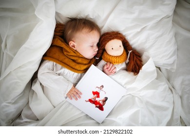 A Cute Caucasian Baby With A Doll Sleeping In Bed With Photo Book , A Sick Child Lies In Bed With A Toy, Bed Rest During Illness. Comfort And Play In Bedroom